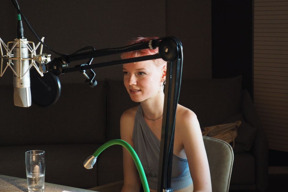 Foto: Rike sitzt an einem Tisch und vor ihr ist ein Mirkrofon angebracht. Sie trägt kurzes pinkes Haar und ein graublaues Oberteil. Ein Anflug eines Lächels liegt auf ihren Lippen.