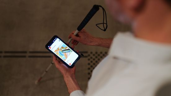 Foto: Eine Person hält ein Audioguide-Gerät und einen Blindenlangstock. Auf dem Boden ist ein taktiler Leitstreifen.