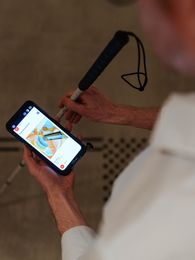 Foto: Eine Person hält ein Audioguide-Gerät und einen Blindenlangstock. Auf dem Boden ist ein taktiler Leitstreifen.