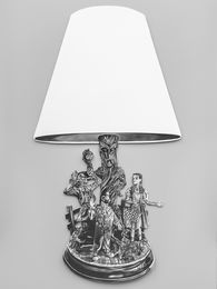 Foto: Nahaufname einer Tischlampe vor grauem Hintergrund, deren unterer Teil aus dunkelgräulich-illustrierten Fabel-Figuren besteht.