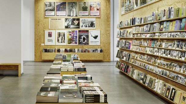 Photo: In the museum shop there are wooden shelves with books, postcards and other products. Posters hang on the wall.