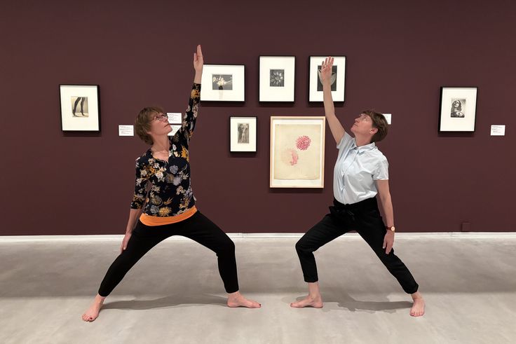 Foto: Zwei Personen in Yoga-Kleidung stehen in jeweils einer Yoga-Position vor gerahmten Fotografien der Künstlerin Marta Astfalck-Vietz.