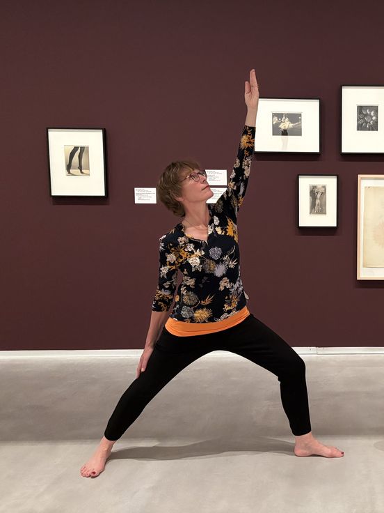 Foto: Zwei Personen in Yoga-Kleidung stehen in jeweils einer Yoga-Position vor gerahmten Fotografien der Künstlerin Marta Astfalck-Vietz.