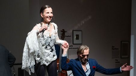 Foto: Meret Becker singt auf einem roten Gymnastik-Ball, während sie von einer weiteren Person gestützt wird.