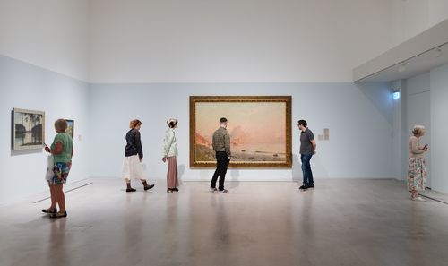 Foto: Einige Personen sehen sich in einem Ausstellungsraum an der Wand hängende Gemälde an.