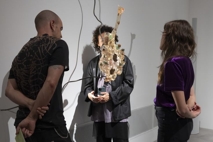Eindrücke aus der Ausstellung „Mariechen Danz. edge out. GASAG Kunstpreis 2024“ Foto: Drei Personen stehen im Halbkreis um ein Kunstwerk von Mariechen Danz, das aus durchlässigem, mehrfarbigem Hartz besteht und an einer dünnen, gebogenen Metall-Strebe befestigt in den Raum hinein ragt.