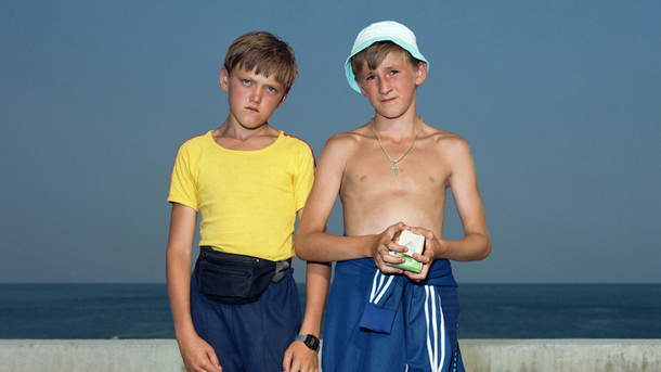 Colour photograph: Two boys stand in front of a concrete balustrade, with the dark blue sea in the background. A cloudless blue sky rises above them. Both boys are wearing tracksuit bottoms and are looking directly into the camera.