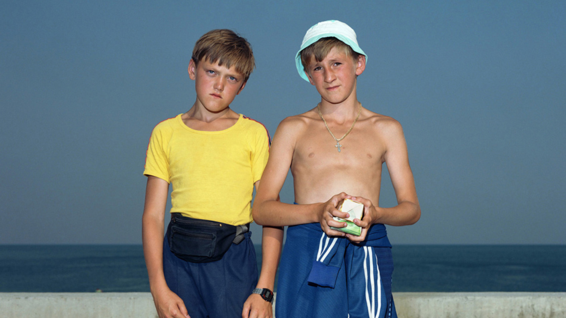 Colour photograph: Two boys stand in front of a concrete balustrade, with the dark blue sea in the background. A cloudless blue sky rises above them. Both boys are wearing tracksuit bottoms and are looking directly into the camera.