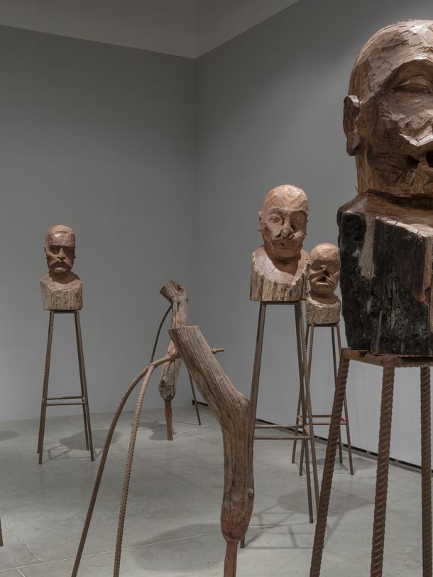 Installation of wooden sculptures standing on pedestals made of fluted metal rods.