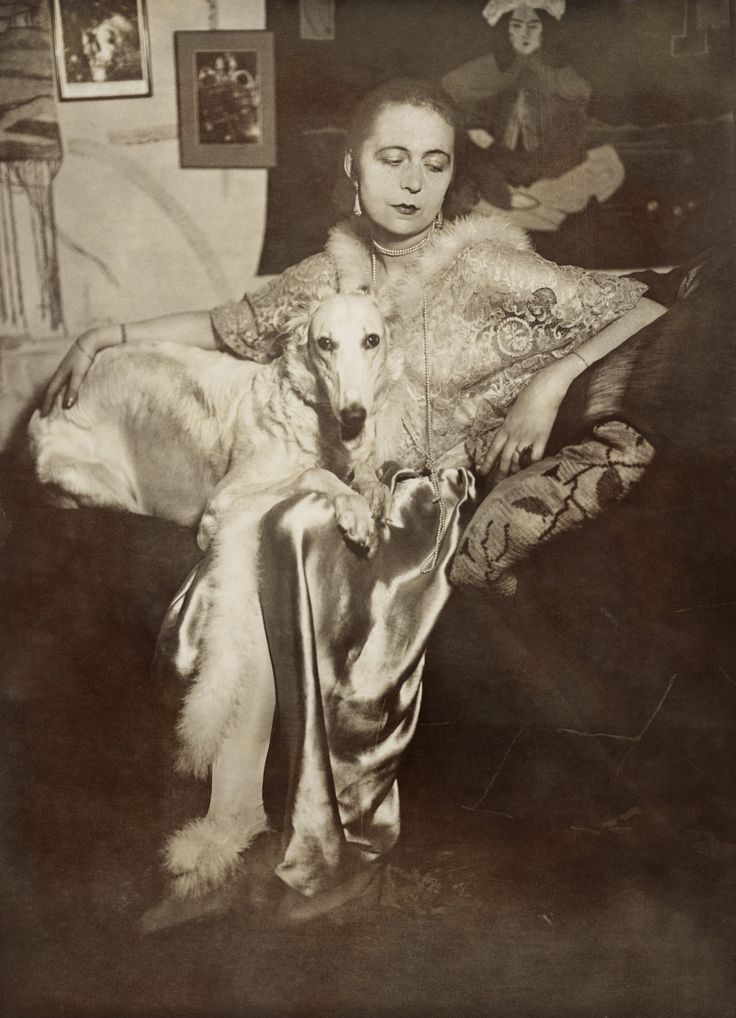 Marta Astfalck-Vietz, Untitled, c. 1927 Self-portrait of the artist Marta-Astfalck-Vietz as a sepia photograph. She is sitting on a sofa in a shiny dress with fur trim. A dog with light-coloured fur sits on her lap. Paintings and decorative elements can be seen in the background.