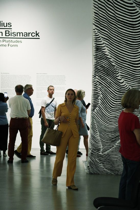 Mehrere Personen stehen im Ausstellungsraum, im Hintergrund ist auf der Wand ein Teil des Ausstellungstitels in schwarzer Schrift angebracht.