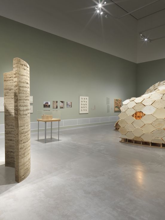 Ausstellungsansicht: Blick auf eine Lehminstallation und eine Konstruktion aus Holz-Pilzmodulen. An den matt-grünen Wänden hängen weitere Arbeiten und Dokumentationen der beteiligten Projekte.