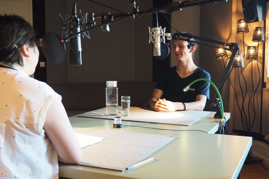 Foto: Elisa und Raphael sitzen sich im Aufnahmestudio gegenüber an zwei Tischen. Vor ihren Köpfen befinden sich Mirofone. Raphael trägt kurzes braunes Haar und ein schwarzes Shirt und spricht.