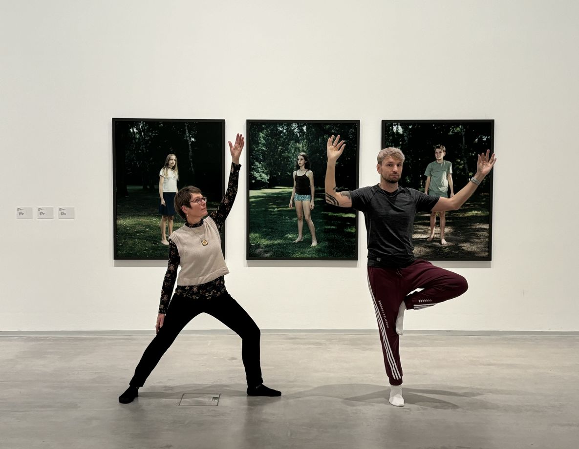 Foto: Zwei Personen in Yoga-Kleidung stehen in jeweils einer Yoga-Position vor drei gerahmten Fotografien der Künstlerin Rineke Dijkstra.