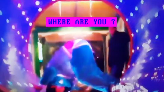 Film still: A person is standing in a near handstand position in a sort of tunnel in bright purple; in the background, another person is standing and looking down at them. The still has a look as if it were taken from a computer game. The words ‘Where are you?’ are also written in bright colours above the person in the tunnel.