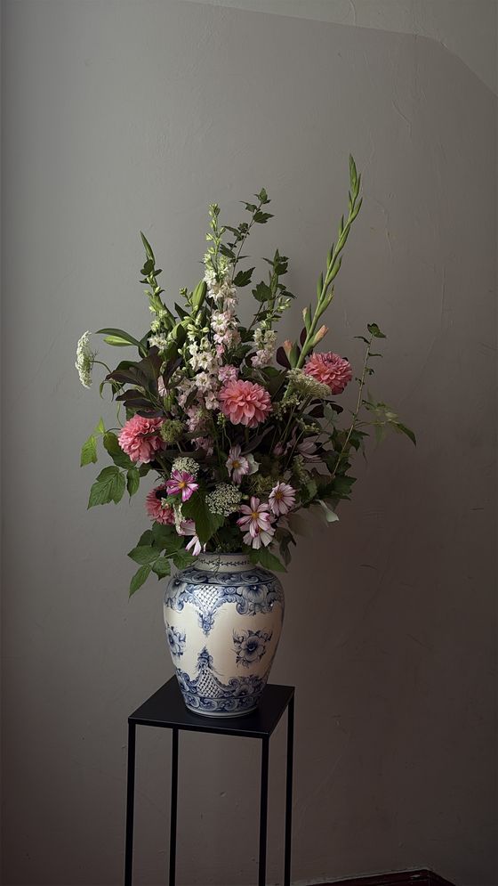 Foto: Ein üppiges Blumen-Bouquet in hellen, pastell-farbenen Tönen steht in einer verzierten Vase vor einer grauen Wand.