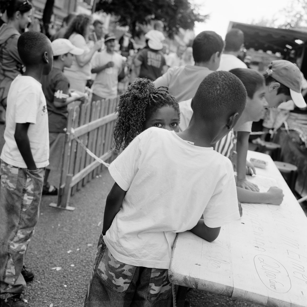 Schwarz-weiß-Fotografie: Blick auf ein Fest im Freien. Zwei Kinder stehen neben einem langen Tisch, eine Person blickt direkt in die Kamera und wird teilweise von der anderen Person verdeckt. 