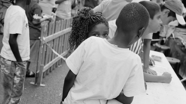 Schwarz-weiß-Fotografie: Blick auf ein Fest im Freien. Zwei Kinder stehen neben einem langen Tisch, eine Person blickt direkt in die Kamera und wird teilweise von der anderen Person verdeckt. 