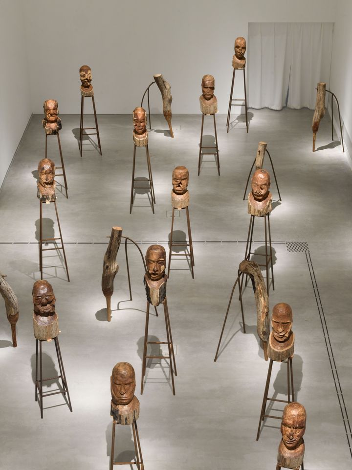 View from above into an exhibition room in which various wooden heads stand on constructions made of grooved metal rods. A white curtain can be seen in the background, leading into the next exhibition room.