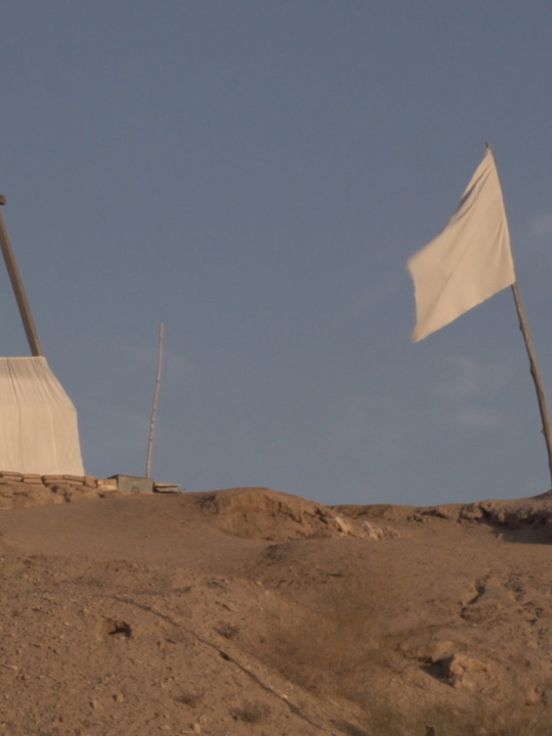 Film still: View of a barren landscape against a mid-blue sky. In the foreground is a white flag on a wooden flagpole, in the background a kind of triangular wooden scaffolding, the lower part of which is covered with white fabric.