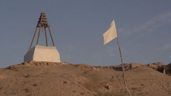 Film still: View of a barren landscape against a mid-blue sky. In the foreground is a white flag on a wooden flagpole, in the background a kind of triangular wooden scaffolding, the lower part of which is covered with white fabric.