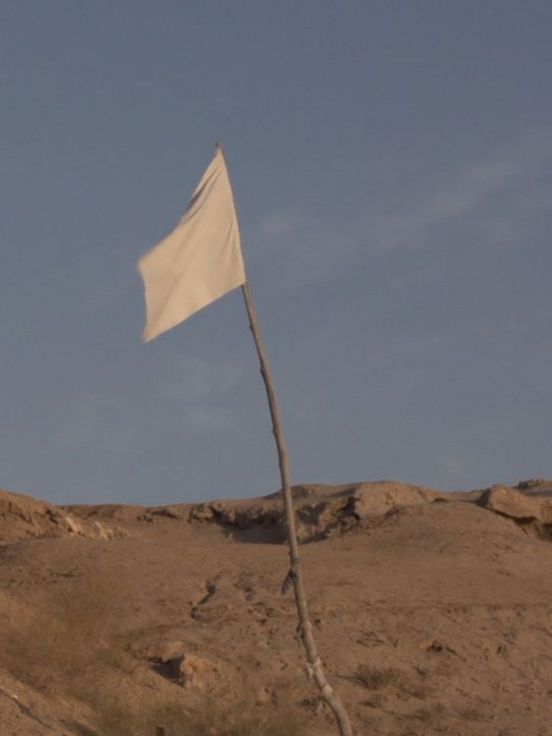 Film still: View of a barren landscape against a mid-blue sky. In the foreground is a white flag on a wooden flagpole, in the background a kind of triangular wooden scaffolding, the lower part of which is covered with white fabric.