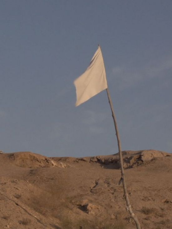 Film still: View of a barren landscape against a mid-blue sky. In the foreground is a white flag on a wooden flagpole, in the background a kind of triangular wooden scaffolding, the lower part of which is covered with white fabric.