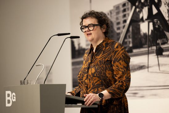 Photo: Ilka Voermann stands at a podium. In the background is a black and white photo wallpaper of a sculpture by Hans Uhlmann at Hansaplatz in Berlin.