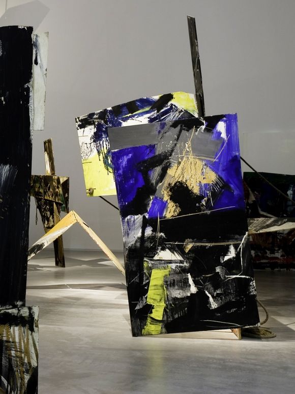 Large-scale abstract installation objects made of painted wooden panels are positioned throughout the exhibition space. The panels feature bold brushstrokes in black, white, grey, yellow, and vibrant blue and are arranged at various angles. The floor is smooth and light, and the walls are neutral with no additional elements.