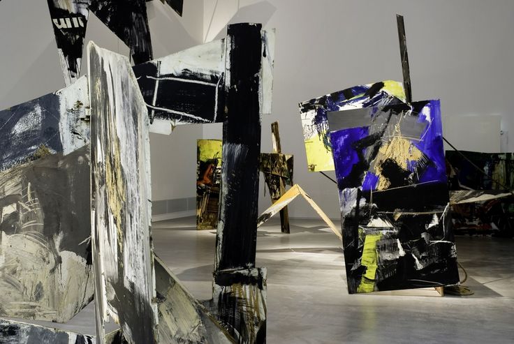 Großformatige, abstrakte Installationsobjekte aus bemalten Holzplatten stehen in einem Ausstellungsraum. Die Platten sind mit kräftigen Pinselstrichen in Schwarz, Weiß, Grau, Gelb und intensivem Blau gestaltet und in verschiedenen Winkeln im Raum positioniert. Der Boden ist glatt und hell, die Wände neutral und ohne zusätzliche Elemente.