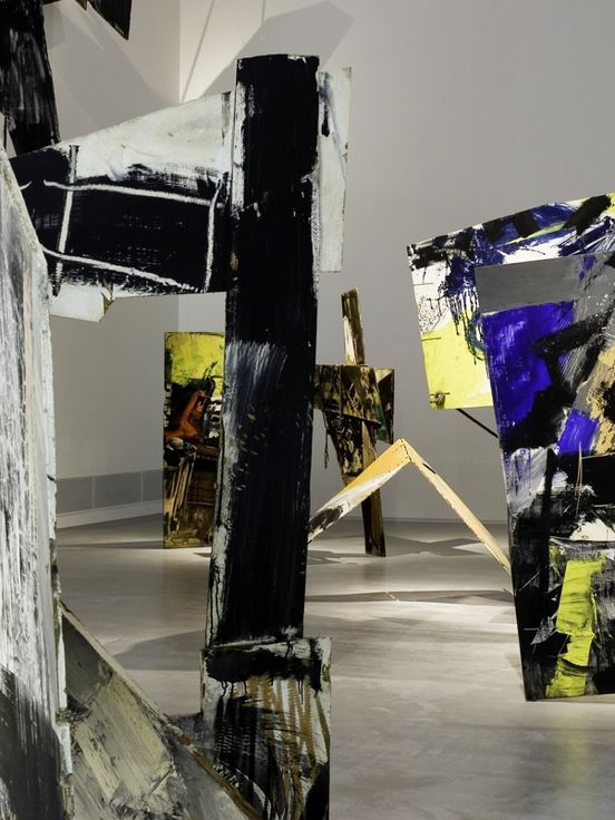 Large-scale abstract installation objects made of painted wooden panels are positioned throughout the exhibition space. The panels feature bold brushstrokes in black, white, grey, yellow, and vibrant blue and are arranged at various angles. The floor is smooth and light, and the walls are neutral with no additional elements.