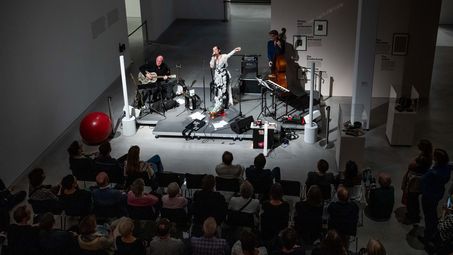 Foto: Die drei Musiker*innen Meret Becker, Yoyo Röhm und Gary Schmalzl auf der Bühne, vor der ein Publikum sitzt.
