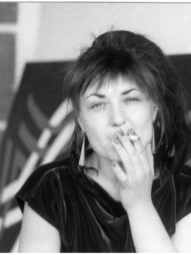 Black and white photo of the artist Käthe Kruse, who is taking a drag on a cigarette and looking directly into the camera. The background shows a door and a painted canvas.