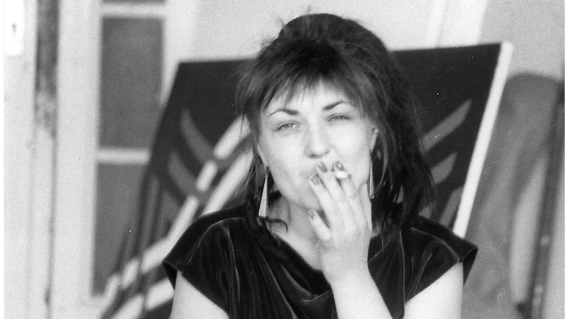 Black and white photo of the artist Käthe Kruse, who is taking a drag on a cigarette and looking directly into the camera. The background shows a door and a painted canvas.