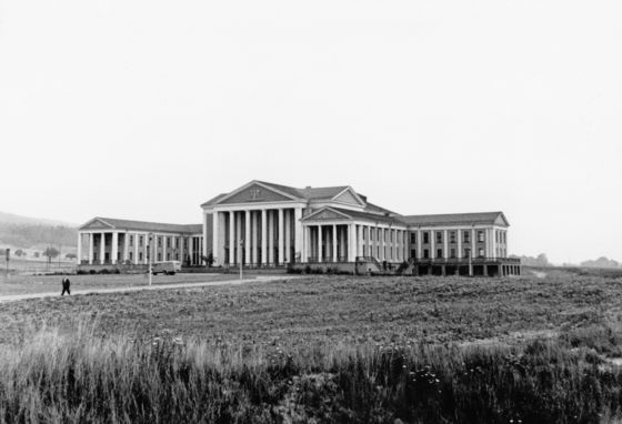 Großes Gebäude mit Säulen und flachen Giebeln inmtten einer Wiese