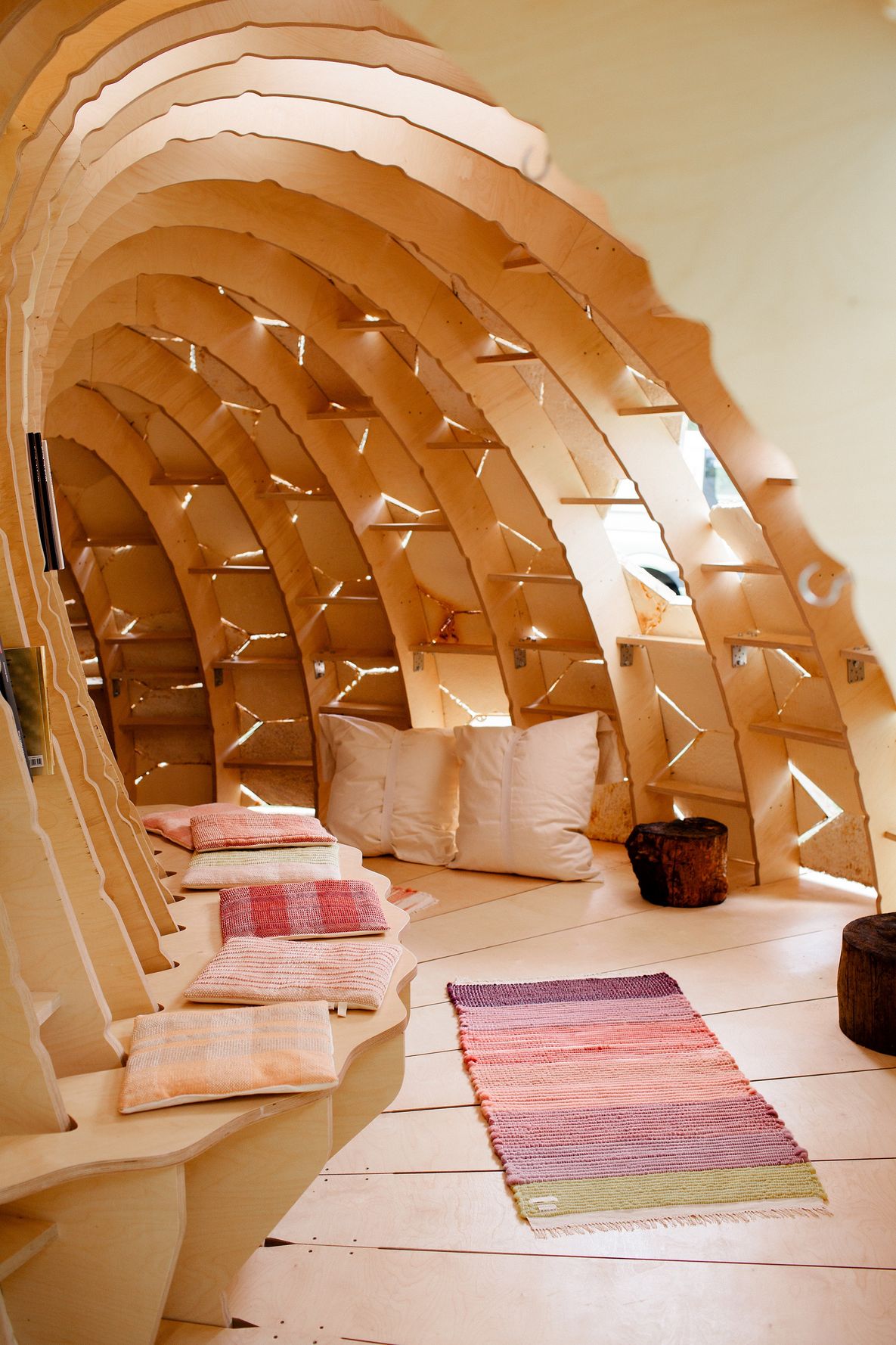 Foto: Blick in einen höhlenförmigen, halbrunden Bau aus Holz mit Sitzbank, einem Teppich und Kissen. Von außen strömt durch Spalten Tageslicht nach innen.