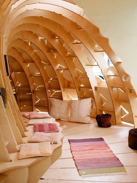 Foto: Blick in einen höhlenförmigen, halbrunden Bau aus Holz mit Sitzbank, einem Teppich und Kissen. Von außen strömt durch Spalten Tageslicht nach innen.