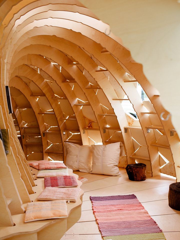 Photo: View into a cave-shaped, semi-circular wooden building with a bench, a carpet and cushions. Daylight streams in from the outside through gaps.