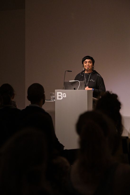  Photo: Monira Al Qadiri stands in front of an audience at a lectern.