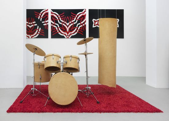 Photo: A leather-covered drum kit stands on a bright red, fluffy carpet. Next to it, a leather banner hangs from the ceiling. In the background is a three-part painting with a red and white graphic pattern on a black background.