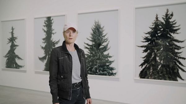 Video still: A person in a pink cap and leather jacket stands in front of a series of photos showing conifers and looks directly into the camera.