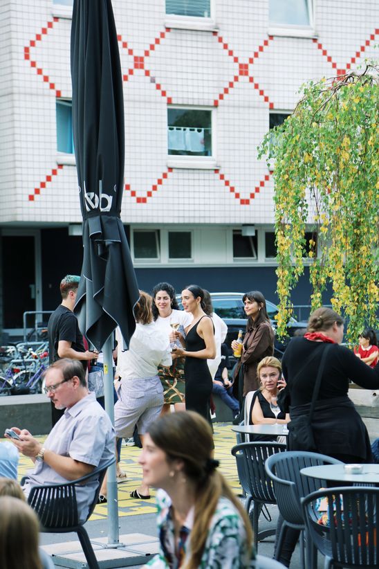 Einige Personen sitzen draußen an Café-Tischen oder stehen, im Hintergrund ein Gebäude mit weiß-roter Fliesenfassade.