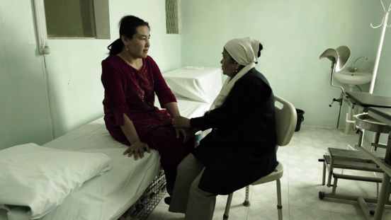 Film still: Two people sitting in a medical treatment room. One person sits on a couch, while the other sits on a chair to the right and holds her hand.