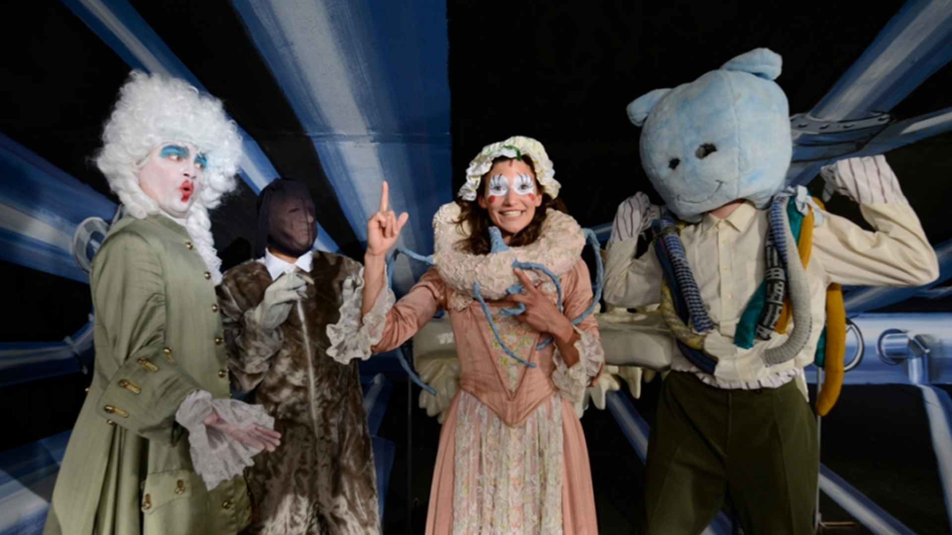 Video still: A group of colourfully costumed people stand next to each other, some of them looking directly into the camera. Two faces are made up like dolls or clowns, while the other two people are each wearing a mask and a plush bear's head.