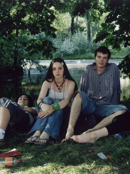 Fotografie: Vier junge Personen sitzen bzw. liegen in Sommerkleidung in einem Park. Ihr direkt ist direkt in die Kamera gerichtet.