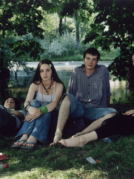 Fotografie: Vier junge Personen sitzen bzw. liegen in Sommerkleidung in einem Park. Ihr direkt ist direkt in die Kamera gerichtet.