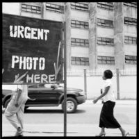 Black and white photograph: The photograph shows two people walking along a road. The upper body of the person in front is covered by a sign that reads “Urgent Photo here”. The background of the picture shows a large building with shaft-like window openings framed by a white fence.