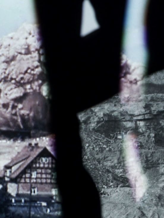 Filmstill: Zweigeteiltes Bild, das in der linken Bildhälfte eine alte, leicht verwackelte und verblichene Aufnahme eines Hauses und einer großen Giftwolke und in der rechten Bildhälfte eine Felsenwand zeigt.