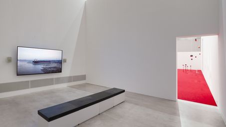 Foto: Blick auf einen Videoscreen und einer Sitzbank, im Hintergrund ist ein Durchgang zum Hauptraum der Ausstellung zu sehen.