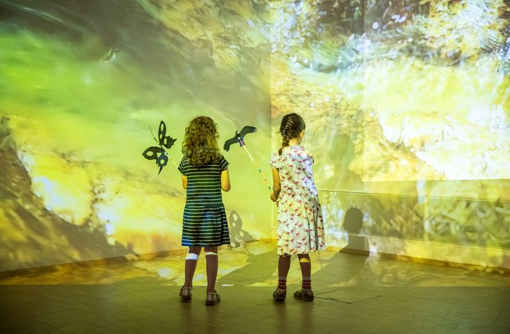 Foto: Rückenansicht von zwei Kindern, die vor einem Licht und Schattenspiel aus gelb-grünlichen Farben mit zwei Schmetterlingen stehen, die an die Wand projiziert werden.